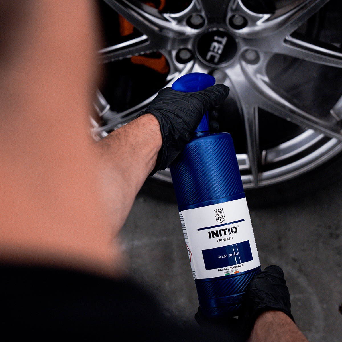 Person holding a bottle labeled &#39;INITIO&#39; near a car wheel