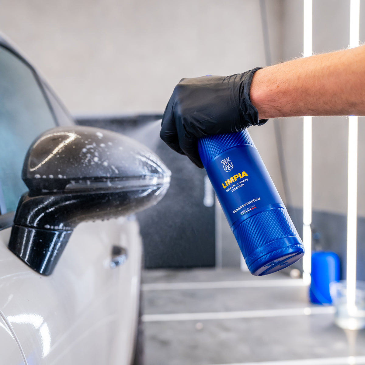 Person cleaning car mirror with a blue bottle labeled &#39;LIMPIA&#39; in a garage setting.