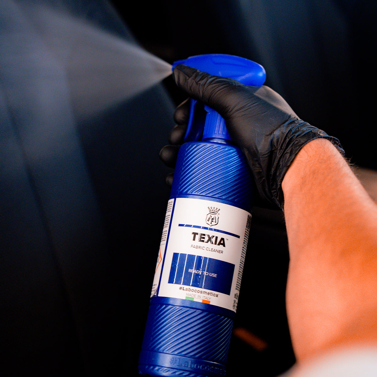 Hand holding a blue spray bottle labeled &#39;TEXIA&#39; against a dark background